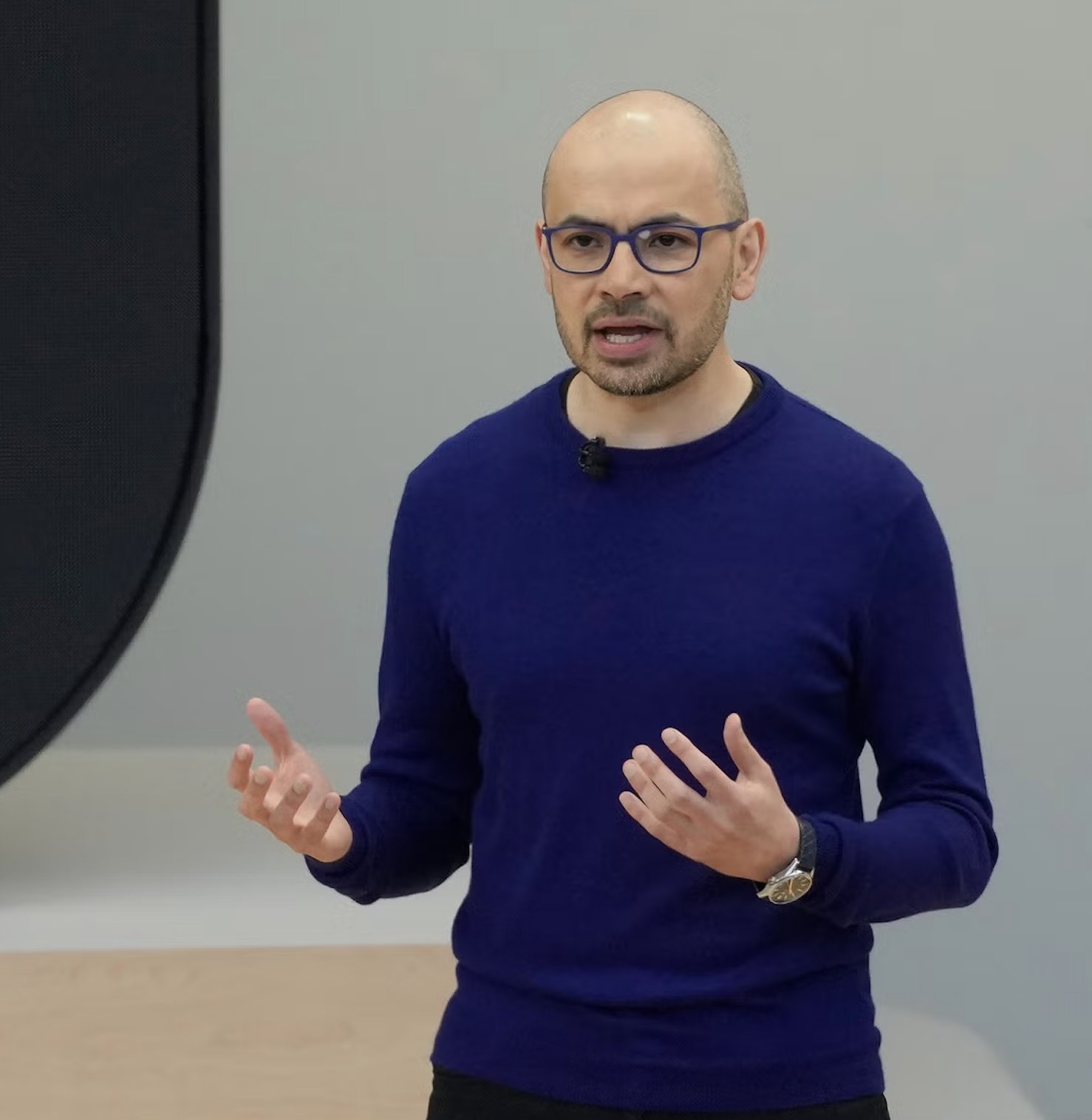 Demis Hassabis 발표 장면 — 파란 스웨터 차림으로 단상에서 <em loading=